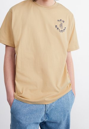 T-shirt en coton couleur crème, à manches courtes, avec un col rond et un petit logo navy, portant le texte "New Balance" et un graphique.