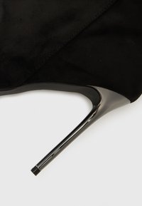 Zapato de tacón alto de ante negro con punta puntiaguda y tacón de aguja brillante. La textura es suave y lisa, con detalles mínimos de costura.