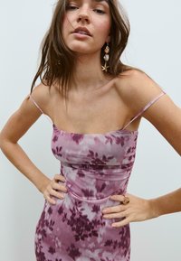 Robe tie-dye violette avec des bretelles fines, design ajusté, texture lisse et motifs floraux subtils. Mannequin portant des boucles d'oreilles accrocheuses et une bague.