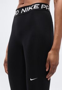 Leggings Nike Pro noirs avec une large ceinture élastiquée et marquée. Tissu lisse et extensible avec un petit logo Nike blanc près du bas.
