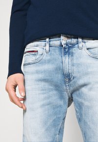 Ljusblå jeans med blekt utseende, femfickdesign och subtila röda och vita detaljer på vänster ficka. Bärs med en marinblå tröja.