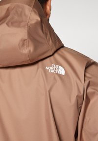 Brun vandtæt jakke med hætte, glat overflade og lille hvidt logo på ryggen. Minimalistisk design uden yderligere detaljer.