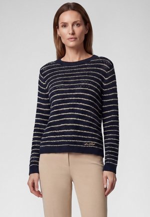 Woman wearing a navy blue and white striped sweater with beige pants, standing against a plain light gray background.
