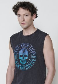 Ärmlös svart t-shirt med en rund grafik som föreställer ett blått skalle och blixt, textil med struktur och avslappnad design.