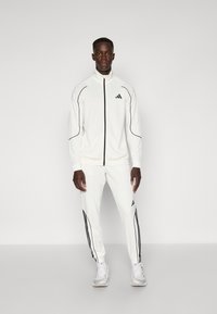 Hvid atletisk sæt med sorte accenter, der inkluderer en lynlåsjakke og matchende bukser, lavet af glat stof, med slim fit og elastiske manchetter.
