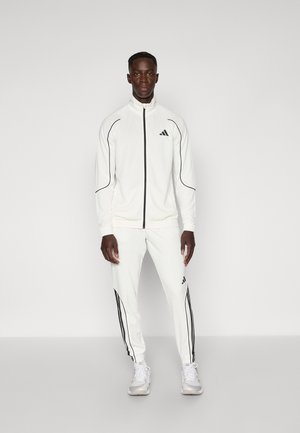 Costum athletic alb cu accente negre, format dintr-o jachetă cu fermoar și pantaloni asortați, realizat dintr-un material neted, cu o croială ajustată și manșete elastice.