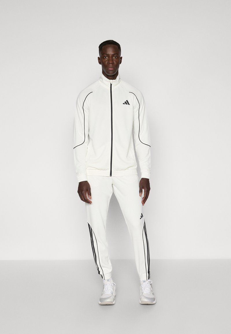 Hvid atletisk sæt med sorte accenter, der inkluderer en lynlåsjakke og matchende bukser, lavet af glat stof, med slim fit og elastiske manchetter.