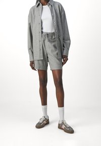 Chemise grise à boutons sur un t-shirt blanc, associée à un short gris. La tenue présente des textures discrètes et est complétée par des chaussettes claires et des baskets brunes.