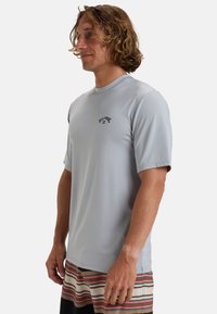 Camisa corta de manga con cuello redondo en gris claro y estampado negro en el pecho, combinada con pantalones cortos a rayas en tonos de beige, naranja y negro.