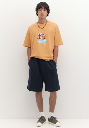 Jeune homme portant un T-shirt beige avec un motif de rouleau à la cannelle, un short bleu marine, des chaussettes blanches et des baskets vertes, debout contre un mur uni.