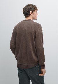 Young man wearing a brown sweater and dark jeans, viewed from behind, standing against a plain light gray background.