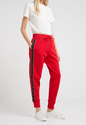 Femme portant un pantalon de survêtement rouge avec des bandes latérales noires « Polo », des baskets blanches et un T-shirt blanc uni à manches courtes, debout devant un fond blanc.