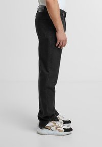 Jeans negri din denim cu tăietură dreaptă, având două buzunare laterale și un buzunar la spate. Purtați cu adidași bej și negri.