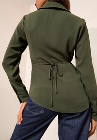 Bluza verde măsliniu cu talie conică și detaliu cu funda, având mâneci lungi și tiv rotunjit, asortată cu blugi albaștri.