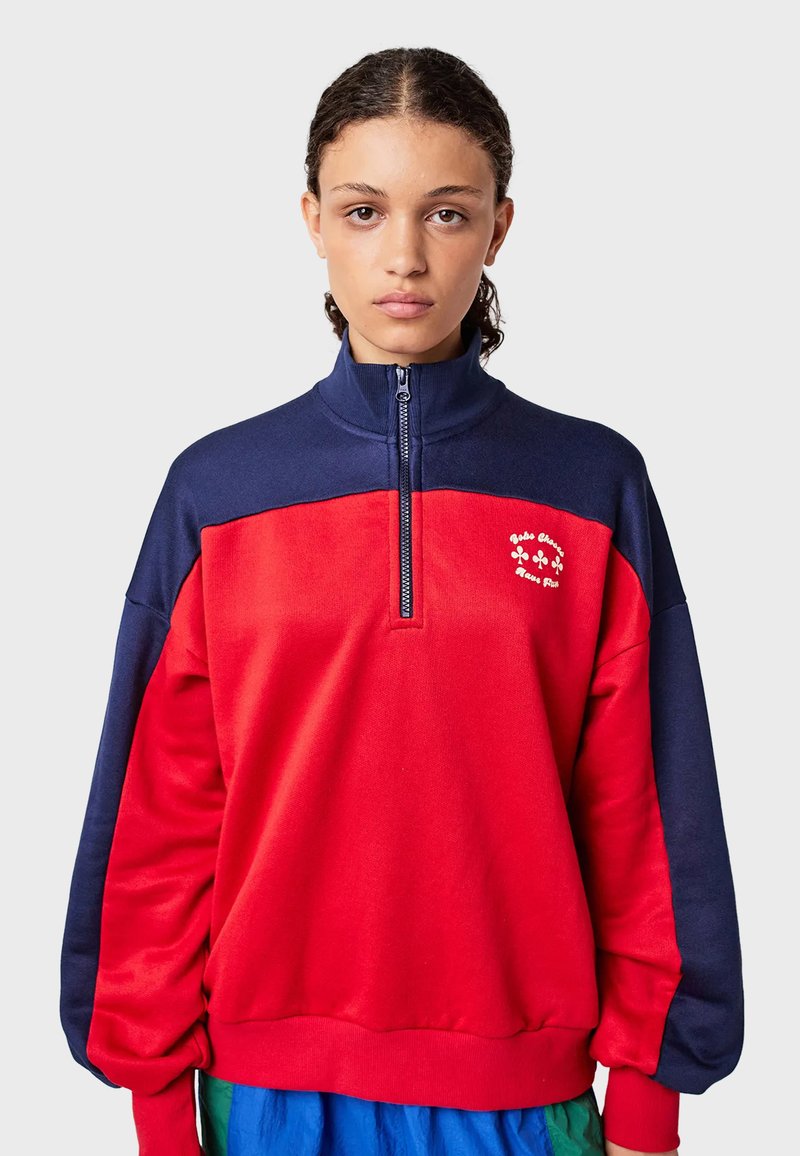 Young person with curly hair wears red and navy quarter-zip sweatshirt with small white clover logo on chest against plain background.