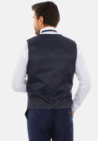 Gilet blu navy con una texture liscia, dotato di chiusura a gancio e occhiello e tracolla posteriore regolabile. Indossato sopra una camicia bianca a maniche lunghe.
