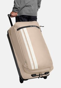 Beige koffer met wielen en witte strepen, gemaakt van duurzaam materiaal. Voorzien van een handvat aan de bovenkant, een ritsluiting en een stevige zwarte basis met wielen.