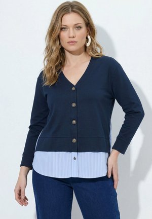 Kvinde med blondt bølget hår iført store hoop-øreringe, en marineblå knappet cardigan over en blåstribet skjorte og mørkeblå jeans, stående mod en hvid væg.