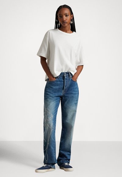 Balta oversized t-krekls kopā ar zilām džinsa biksēm, kurām kreisajā kājā ir gaiši zils izšuvums; papildināts ar zilām sporta kurpēm.