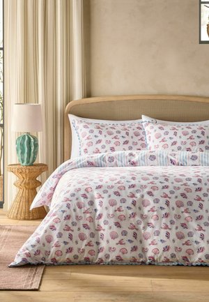 Bett mit beigem gewebtem Kopfteil, weißer Bettwäsche mit roten und blauen Muschel- und Hummermustern, Rattanseitentisch mit grünem Lampenschirm und beigen Vorhängen.