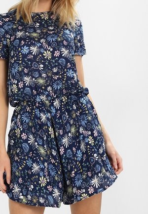Kvinde iført en mørkeblå kortærmet romper med et flerfarvet blomsterprint og en elastisk talje med bindebælte.