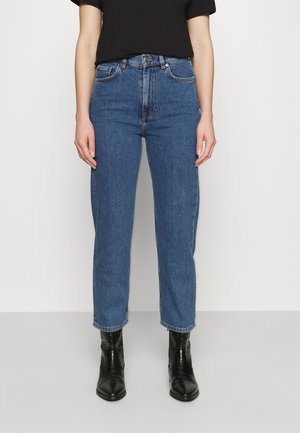 Person iført blå jeans med høj talje, sort t-shirt med korte ærmer og sorte ankelstøvler, står mod en ensfarvet lys baggrund.