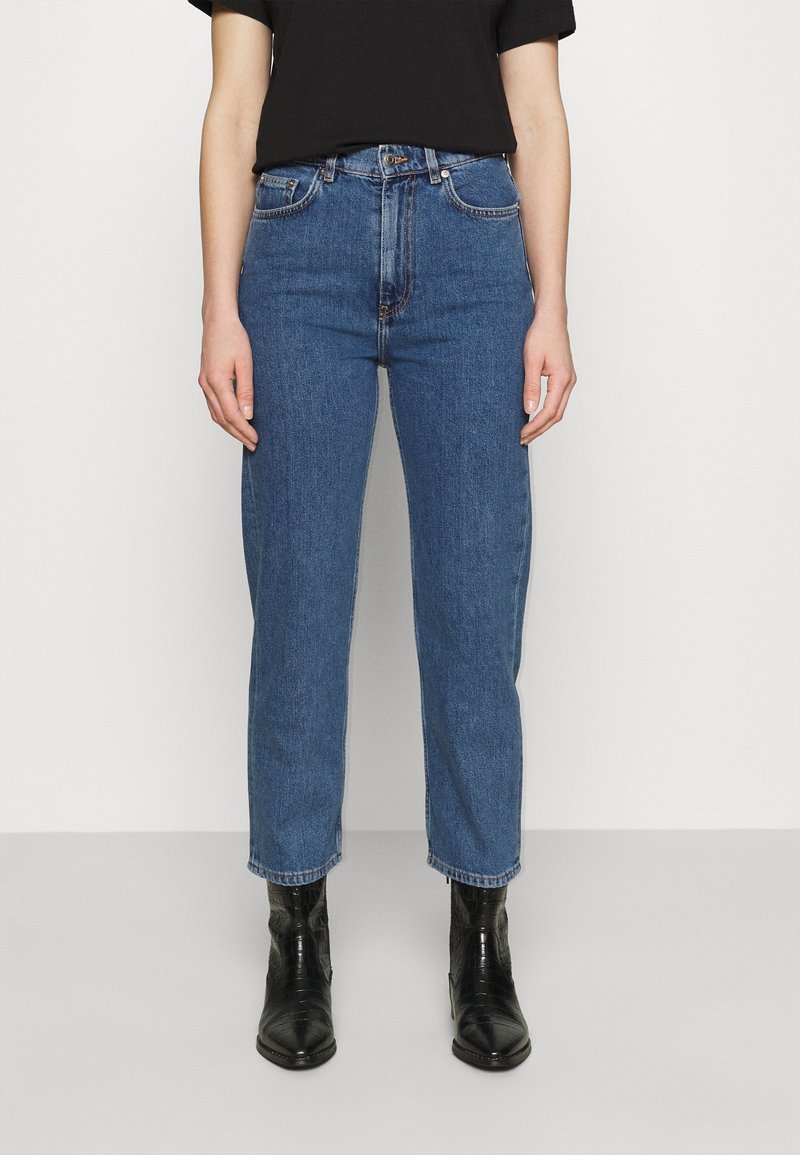 Personne portant un jean bleu taille haute, un t-shirt noir à manches courtes et des bottines noires, debout devant un fond clair uni.
