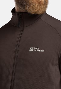 Veste zippée marron en tissu texturé, dotée d'un col montant et d'un logo blanc sur la poitrine, conçue pour les activités de plein air.