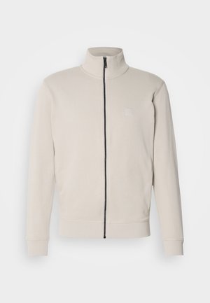 Lys beige zip-up jakke med høy krage, ribbestrikkede mansjetter og en liten logo på brystet. Myk stoff og strømlinjeformet design.