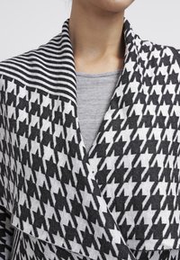 Černobílý kabátek s vzorem houndstooth a pruhovaným límcem, vyrobený z měkké látky. Obsahuje vrstvený přední design a elegantní límec.