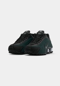 Um par de sapatilhas Nike Shox pretas e verde-escuro com amortecimento em mola visível nos calcanhares, posicionadas lado a lado sobre um fundo branco.