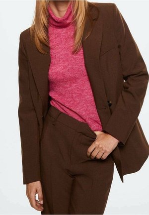 Osoba ubrana w brązowy garnitur z dopasowanymi spodniami i różowym swetrem z golfem, z jedną ręką w kieszeni spodni.
