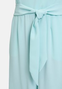 Ljust blå klänning i ett texturerat tyg, med ett knutet bälte vid midjan och mjukt drapering som skapar en elegant silhuett.