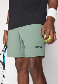 Shorts de sport verts avec le logo "BOSS" noir sur le côté, fabriqués en tissu lisse. Main tenant une balle de tennis jaune et une raquette.