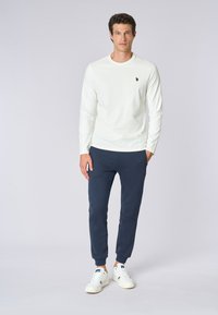 Camicia bianca a maniche lunghe con un piccolo logo, abbinata a pantaloni jogger blu navy e scarpe da ginnastica bianche. Design semplice, tessuto in cotone, vestibilità comoda.