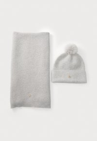 Lichtgrijze gebreide sjaal naast een bijpassende beanie, met een pompon en een klein goudkleurig logo op de boord. Zachte textuur, eenvoudig ontwerp.