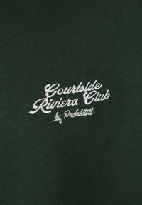 Texte blanc brodé « Courtside Riviera Club by Punchlisted » sur fond de tissu vert foncé.
