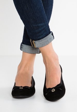 Fødder iført sorte balletsko med små sløjfer og runde metalliske vedhæng, parret med opkrogede mørkeblå jeans.