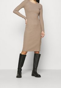Robe beige à manches longues côtelée avec une silhouette ajustée, ourlet jusqu'aux genoux, associée à des bottes en cuir noir hautes avec une finition texturée.