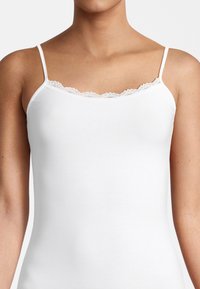 Camisola bianca a costine con spalline sottili e un bordo in pizzo lungo il colletto, caratterizzata da un design aderente e una superficie liscia.