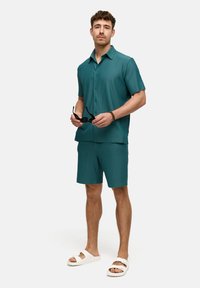 Türkise kurzärmelige Rippbluse und passende Shorts, kombiniert mit weißen Sandalen. Das Model hält eine Sonnenbrille und zeigt ein entspannt geschnittenes Design.