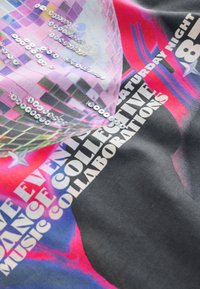 Graphic t-shirt featuring a multi-colored print with sequins. Design includes text, patterns, and a mix of pink, black, and gray hues.