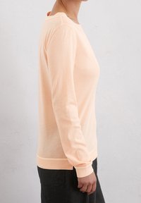 Helles Pfirsich langärmliges Sweater mit Rundhalsausschnitt, gerippten Bündchen und Saum, aus weichem, strukturiertem Stoff, getragen über dunklen Hosen.