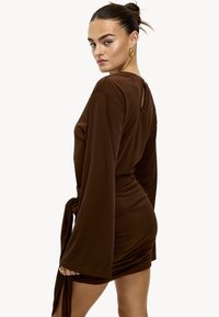 Robe portefeuille marron en tissu doux, avec manches longues et larges, détail dos en forme de goutte, et taille nouée, portée en version mini.