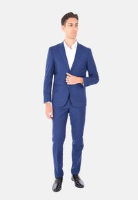 Abito blu navy realizzato in tessuto leggero, design strutturato, vestibilità slim, abbinato a una camicia bianca e scarpe stringate nere.