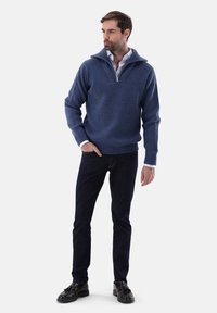 Blauer gerippter Strickpullover mit einem halben Reißverschlusskragen, über einem weißen Hemd mit Knöpfen getragen, kombiniert mit dunkelblauen Jeans und schwarzen Schuhen.
