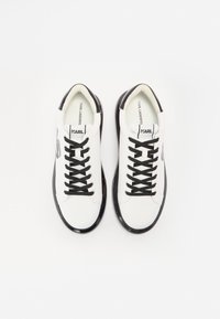 White sneakers with black accents, featuring a smooth leather-like upper, black laces, and rounded toe. Logo printed inside.