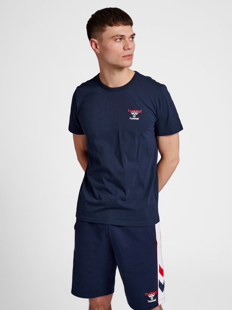 Jonge man in marineblauw hummel sportkleding, kijkt naar links met handen achter de rug tegen een effen witte achtergrond.