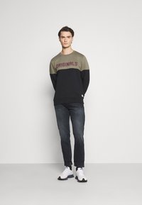 Sweatshirt preto e verde oliva com a palavra "ORIGINALS" em bordô. Combinado com jeans escuros e ténis brancos. Textura suave, corte casual.