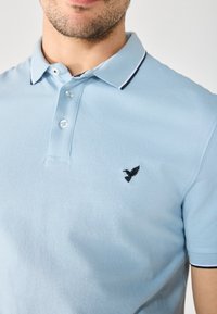 Svijetloplava polo majica od pamuka, s ovratnikom, tri gumba na prednjem dijelu i crnim izvezenim logom ptice na prsima.